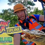 Zipline guide in orange helmet and safety vest poses beside a large sign for Screamin' Gator Zipline at Gatorland, Orlando, Florida with an alligator nearby.