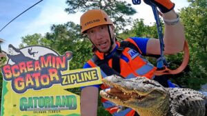 Zipline guide in orange helmet and safety vest poses beside a large sign for Screamin' Gator Zipline at Gatorland, Orlando, Florida with an alligator nearby.