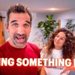 Two people in a bright living room; man in red speaks to the camera while a woman with curly hair watches, with 'TRYING SOMETHING NEW' text overlay.