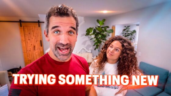 Two people in a bright living room; man in red speaks to the camera while a woman with curly hair watches, with 'TRYING SOMETHING NEW' text overlay.