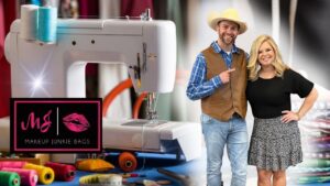 Sewing machine on a blue-worktable with colorful thread spools and fabric; logo reads 'Makeup Junkie Bags' as two hosts stand nearby, smiling.