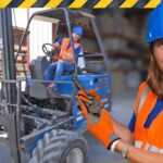 Warehouse scene with a forklift and two workers in safety gear; foreground worker smiles at the camera in an orange vest and blue helmet.