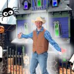 Man in a cowboy hat and vest poses with arms spread in front of a Halloween haunted house decorated with cartoon ghosts and carved pumpkins.