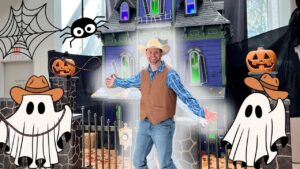 Man in a cowboy hat and vest poses with arms spread in front of a Halloween haunted house decorated with cartoon ghosts and carved pumpkins.