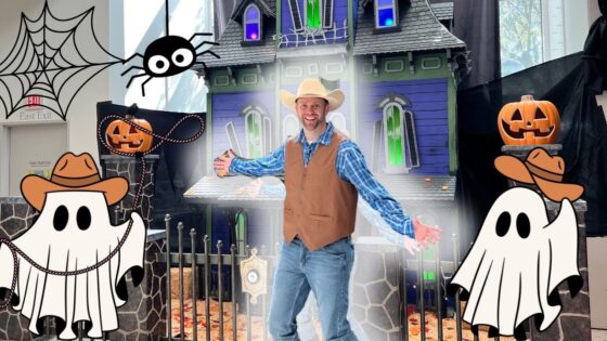 Man in a cowboy hat and vest poses with arms spread in front of a Halloween haunted house decorated with cartoon ghosts and carved pumpkins.
