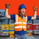 Confident man in a blue hard hat and orange safety vest standing in front of two fire trucks, arms raised in celebration.