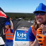 Man wearing a blue hard hat and orange vest with a young girl on the beach, as a large black shark-shaped kite unfolds for Huntington Beach's Kite Festival in the background.
