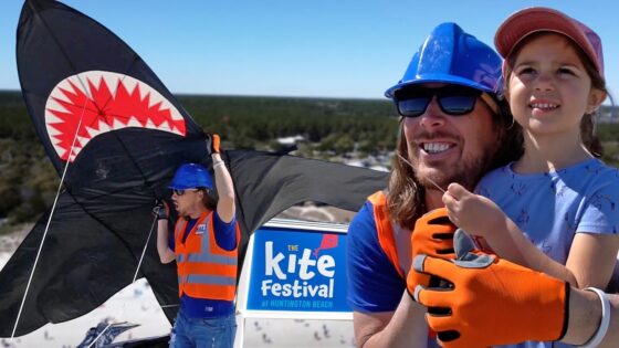 Man wearing a blue hard hat and orange vest with a young girl on the beach, as a large black shark-shaped kite unfolds for Huntington Beach's Kite Festival in the background.