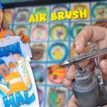 Smiling man in a blue hard hat and orange safety vest holds up a white T‑shirt with a bright airbrushed design, while an airbrush tool is shown in the foreground and a colorful grid backdrop fills the scene.