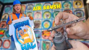 Smiling man in a blue hard hat and orange safety vest holds up a white T‑shirt with a bright airbrushed design, while an airbrush tool is shown in the foreground and a colorful grid backdrop fills the scene.