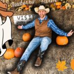 Smiling man in a cowboy outfit sits on hay bales with pumpkins and autumn decor around him, Halloween vibe.