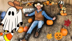 Smiling man in a cowboy outfit sits on hay bales with pumpkins and autumn decor around him, Halloween vibe.