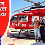 Two men in cowboy outfits pose beside a bright red Life Flight helicopter on a sunny tarmac behind the scene banner. The left shows a bold 'Life Flight Learn About Helicopters!' graphic.