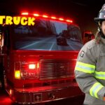 Smiling firefighter in turnout gear with a red fire truck inside a station and bold 'FIRE TRUCK' text in yellow above.
