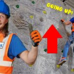 Climber in an orange safety vest and blue helmet ascends a rock-climbing wall, while another worker faces the camera and smiles with a thumbs up; a red arrow and 'GOING UP!' text appear.