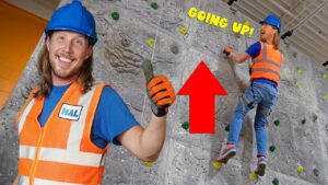 Climber in an orange safety vest and blue helmet ascends a rock-climbing wall, while another worker faces the camera and smiles with a thumbs up; a red arrow and 'GOING UP!' text appear.