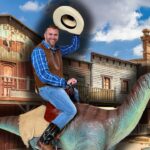Man in a plaid shirt and brown vest rides a dinosaur statue through a Wild West street, tipping his hat cheerfully.