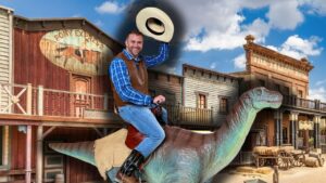 Man in a plaid shirt and brown vest rides a dinosaur statue through a Wild West street, tipping his hat cheerfully.