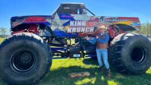 Man in cowboy hat and vest stands next to a giant monster truck named King Krunch on a grassy field.
