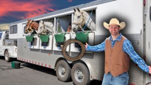Smiling cowboy in a straw hat and brown vest stands beside a horse trailer; horses lean from the stalls with green feed buckets nearby.