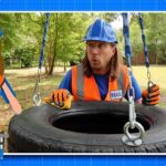 Man in blue hard hat and orange safety vest hoists a tire with a chain in a park; HAL badge visible, with a cartoon HAL character on the left.