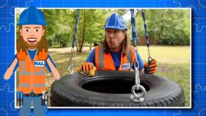 Man in blue hard hat and orange safety vest hoists a tire with a chain in a park; HAL badge visible, with a cartoon HAL character on the left.