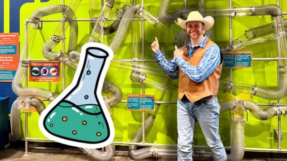 Smiling man in a cowboy hat and brown vest points at a large cartoon flask in a bright lab exhibit with tangled metal tubes behind him.