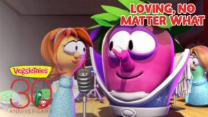 Pink VeggieTales character sings into a microphone while other colorful veggie characters look on on a stage; banner reads 'Loving, no matter what'.