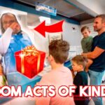 Family of four in line at a store counter looks at a life-size cutout of a man, with a red gift box and Christmas decorations nearby; bold 'RANDOM ACTS OF KINDNESS' caption visible.