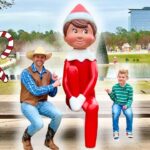 Man in a cowboy hat and vest sits on a bench beside a large red elf statue while a smiling boy sits on the other end, in a Christmas-decorated park.