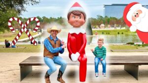Man in a cowboy hat and vest sits on a bench beside a large red elf statue while a smiling boy sits on the other end, in a Christmas-decorated park.