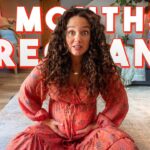 Pregnant woman with curly hair sitting on a living room rug, hands on her belly, with bold '8 MONTHS PREGNANT' overlay.
