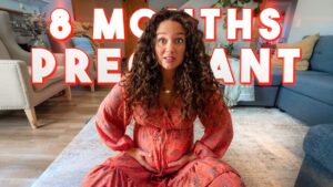 Pregnant woman with curly hair sitting on a living room rug, hands on her belly, with bold '8 MONTHS PREGNANT' overlay.