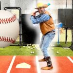 Man in a cowboy hat and vest batting in a indoor cage, swinging at a giant oversized baseball balloon.