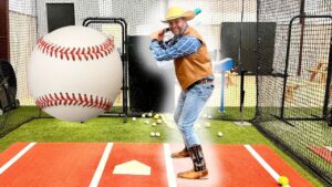 Man in a cowboy hat and vest batting in a indoor cage, swinging at a giant oversized baseball balloon.