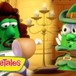 Two green VeggieTales characters sit at a wooden table in a bright castle room; one with a brown wig, the other wearing a green hat with a feather, logo visible in corner.