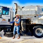 Road crew worker wearing a hat and vest stands beside a white Tymco street-sweeper truck on a sunny road, with orange traffic cones in front.