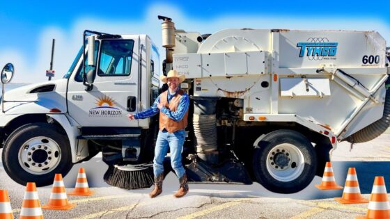 Road crew worker wearing a hat and vest stands beside a white Tymco street-sweeper truck on a sunny road, with orange traffic cones in front.