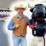 Smiling man in a cowboy hat and brown vest posing with a bull mascot in a bright locker room.