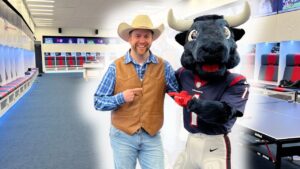 Smiling man in a cowboy hat and brown vest posing with a bull mascot in a bright locker room.