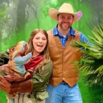 Woman in a green jacket holding a colorful dinosaur puppet, smiling with a man in a brown vest and cowboy hat in a jungle backdrop.