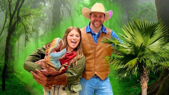 Woman in a green jacket holding a colorful dinosaur puppet, smiling with a man in a brown vest and cowboy hat in a jungle backdrop.