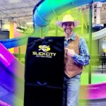 Man in a cowboy hat and vest holds a black Slick City Action Park bag beside colorful indoor slides in the background.