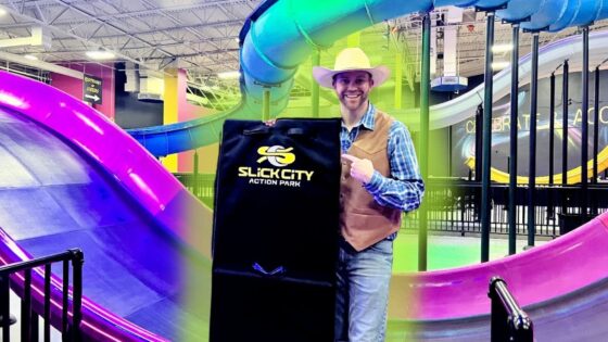 Man in a cowboy hat and vest holds a black Slick City Action Park bag beside colorful indoor slides in the background.