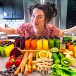 Woman in a kitchen with arms spread wide, presenting a row of colorful bottled juices and fresh produce on a countertop.