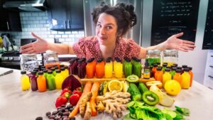 Woman in a kitchen with arms spread wide, presenting a row of colorful bottled juices and fresh produce on a countertop.