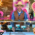 Smiling man in a cowboy hat and brown vest behind a pink, heart-themed ice cream shop counter, with cookies and decorations nearby.