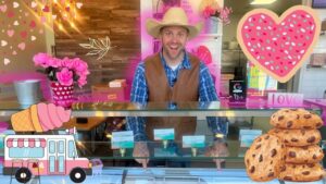 Smiling man in a cowboy hat and brown vest behind a pink, heart-themed ice cream shop counter, with cookies and decorations nearby.