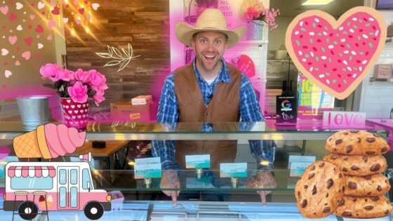 Smiling man in a cowboy hat and brown vest behind a pink, heart-themed ice cream shop counter, with cookies and decorations nearby.