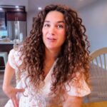 Woman with long curly hair sits at a wooden dining table in a bright kitchen, smiling at the camera. (Informative, concise)